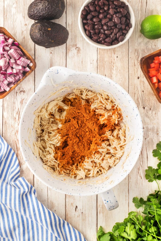 A white bowl filled with shredded chicken and taco seasoning sits on a wooden table, surrounded by bowls of black beans, chopped red onions, diced tomatoes, avocados, fresh cilantro, and a lime.