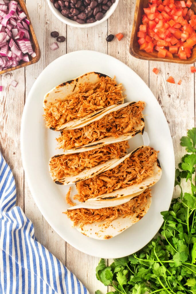 Four shredded chicken tacos on a white oval plate, surrounded by bowls of black beans, diced tomatoes, chopped red onions, fresh cilantro, and a blue striped napkin on a rustic wooden table.