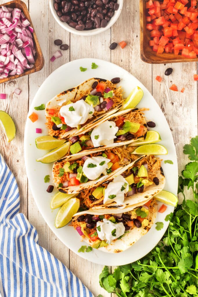 Four chicken tacos topped with chopped tomatoes, red onions, avocado, black beans, cilantro, and sour cream are arranged on a white plate with lime wedges, surrounded by fresh vegetables and herbs on a rustic table.