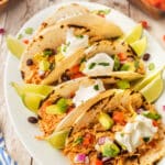 A plate of four soft chicken tacos topped with sour cream, diced tomatoes, red onions, avocado, black beans, and cilantro, garnished with lime wedges. Bowls of chopped vegetables are in the background.