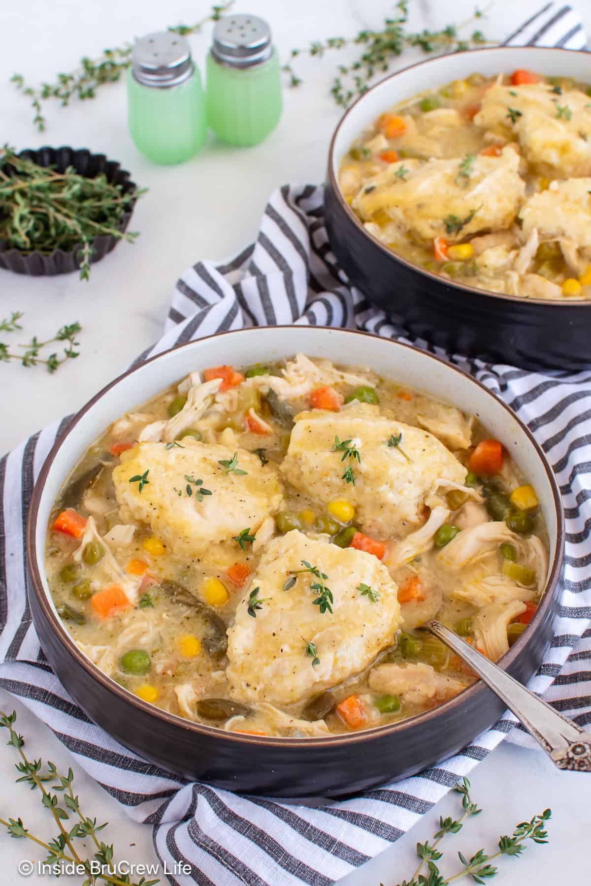 Two bowls of chicken and dumplings soup filled with vegetables like carrots, peas, and corn, topped with fluffy dumplings and garnished with fresh thyme, set on a striped towel with salt and pepper shakers in the background.