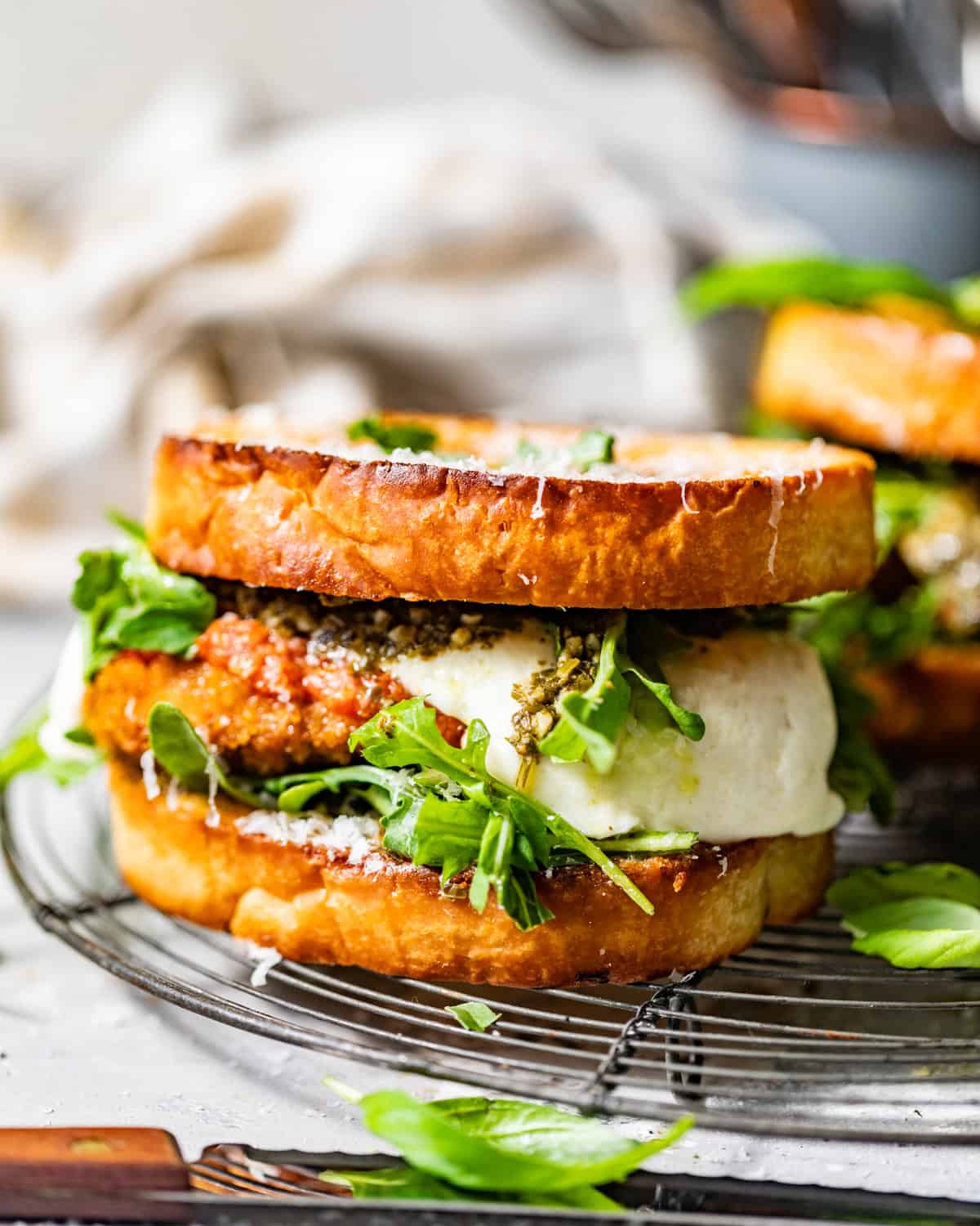 A close-up of a gourmet sandwich on toasted bread, filled with melted cheese, crispy breaded patty, fresh arugula, and pesto, sitting on a wire rack with some greens scattered around.