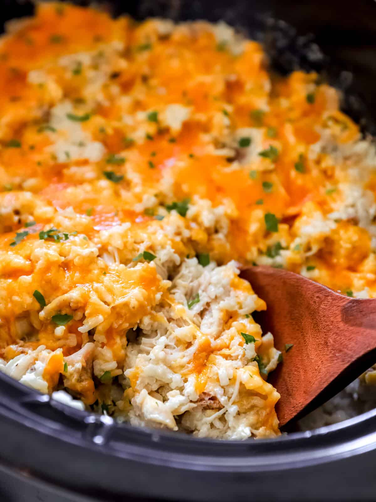 A close-up of a creamy, cheesy casserole with shredded chicken and rice being scooped with a wooden spoon, topped with melted cheddar cheese and chopped herbs.