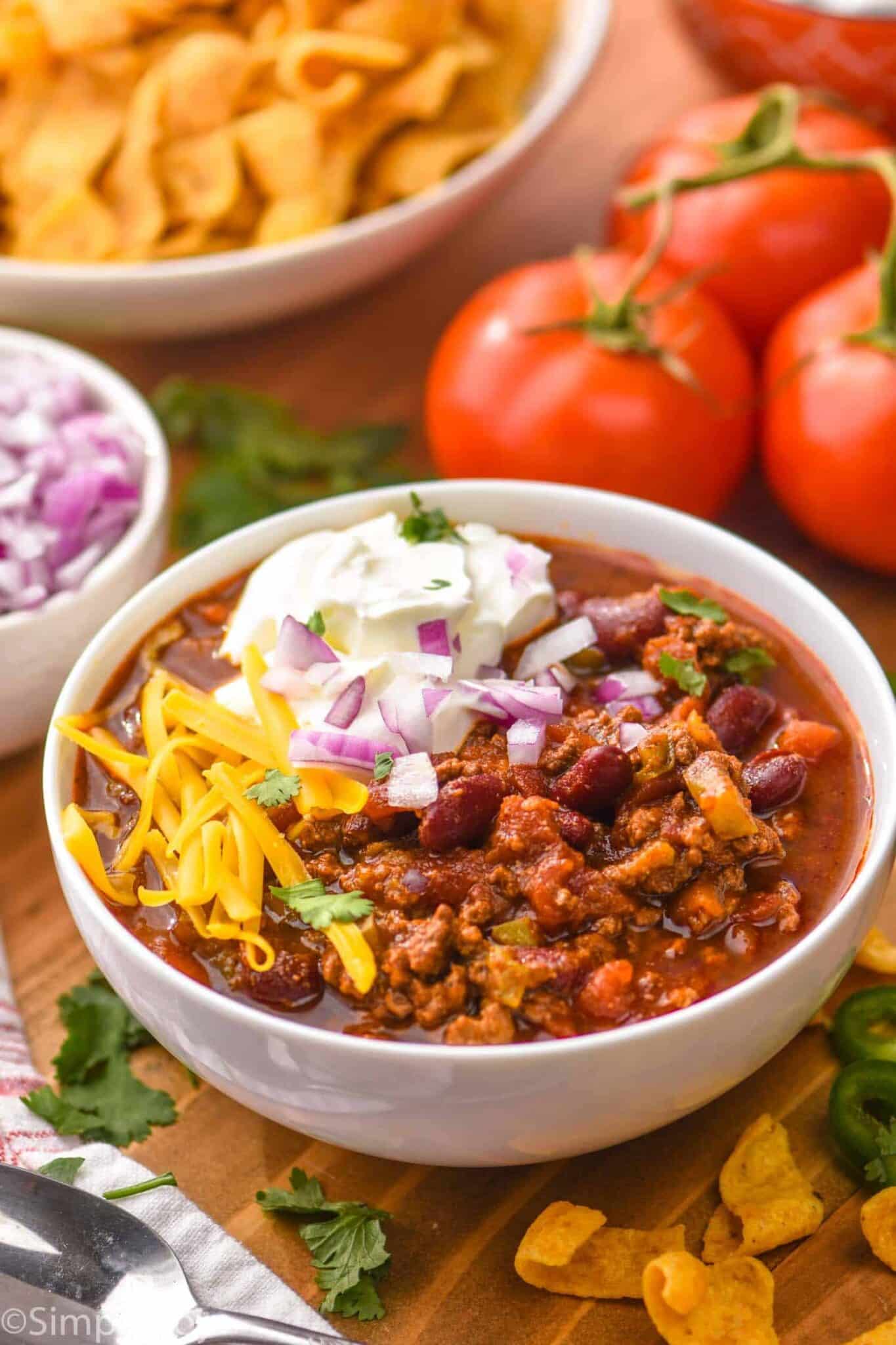 A bowl of chili topped with shredded cheddar cheese, chopped red onions, and a dollop of sour cream, surrounded by fresh tomatoes, a bowl of corn chips, cilantro, and a small bowl of chopped onions.