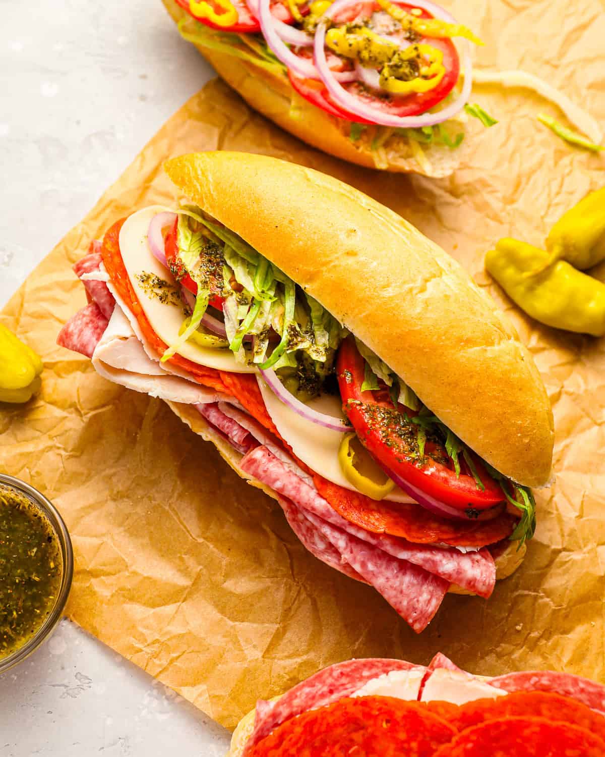 A sub sandwich on parchment paper filled with layers of sliced meats, provolone cheese, lettuce, tomato, onion, and peppers, with Italian dressing drizzled on top. Whole pepperoncini peppers are placed nearby.